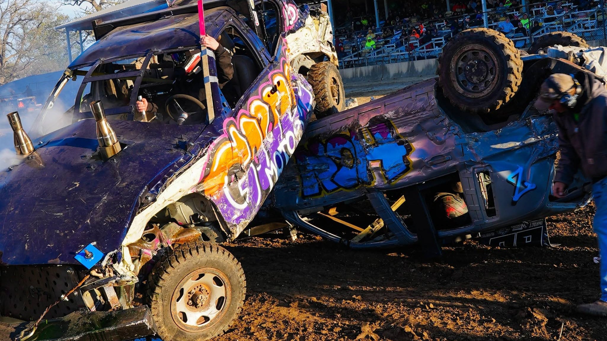 A damaged purple and blue race car with graffiti-style graphics is shown crashing into another vehicle in a muddy outdoor arena, with spectators visible in the background stands.
